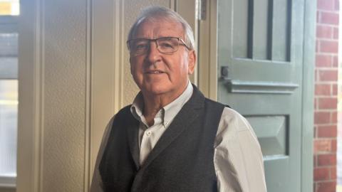 John Armstrong is wearing a white shirt and grey waistcoat. He is wearing glasses and has grey hair. He is smiling at the camera and standing in front of a green corner door of the chronicle press with its exterior red bricks in the background.
