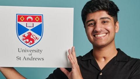 Eshan Bilal smiling, standing and holding a sign with a shield design saying University of St Andrews