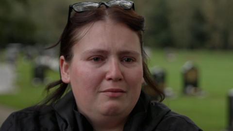 A close-up of Jasmine Beverley during an interview at a cemetery, with gravestones visible in the background.