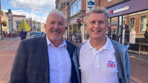 The picture shows two men facing the camera. They are standing in Spalding town centre. On the left, wearing a white shirt and grey jacket, is Julian Wheeler. He has short white hair and a goatee beard. On the right, stands Paul Parkin. He has spiky grey hair and is wearing a white polo shirt and light grey windcheater. 