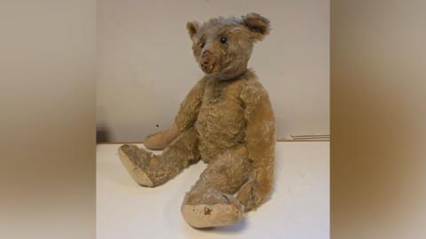 A light brown teddy bear, showing clear signs of use and missing fur. It is sitting upright on a white table.
