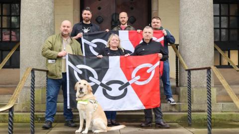 A group of six people are standing on stone steps leading up to large wooden doors. There are handrails either side. Three men are standing in the back row holding a large black, white and red flag with a chain link design on it. There are two men and one woman, who is standing in the middle, in the row in front. They are also holding a flag with the same details on it. There is a large guide dog in front of them. The people in the image are: Karl Denning, Steve Edwards, Karen Westwood, Peter Drake, Pete Lowe, Matt Cook.