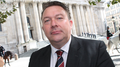 A man with short dark hair, a black suit jacket, red and black tie, and white shirt, stands outside the front of a large building with white pillars.