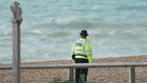 A police community support official stands on the beach, looking towards the sea