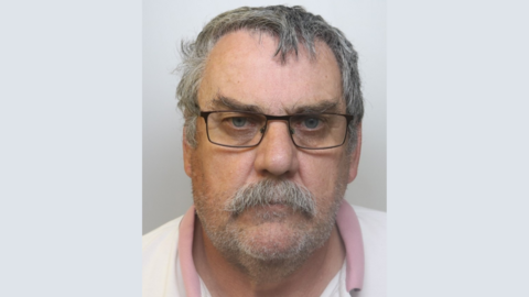 A police mugshot of Philip Garrett, wearing a white shirt with a pink collar and black rectangular glasses. He has dark grey hair and a grey moustache, and is standing against a grey background looking at the camera with a blank expression.