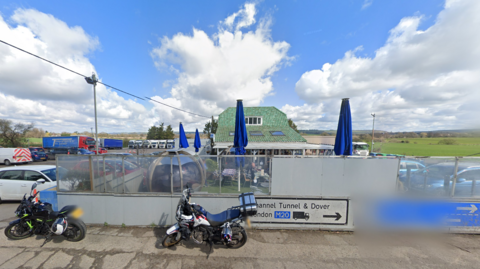 The exterior of the Airport Cafe in Sellindge, on a sunny day, with tables and chairs and the sign showing opening times.