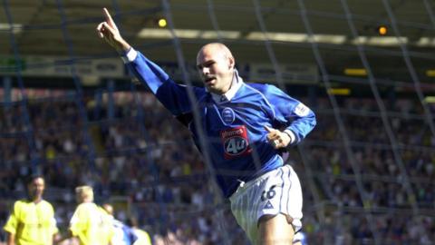 Tommy Mooney holds his right arm up and points his finger up to the sky as he runs. he wears a long sleeve blue shirt and white shorts