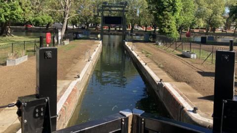 Bedford Lock, showing black large gates, at one end, and a green lock at the other, with a water channel in the middle and The Embankment in the distance, with trees along it. The water is dark. 