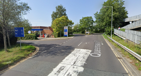 A road leading to a Tesco petrol station. There are green trees and the sky is blue.
