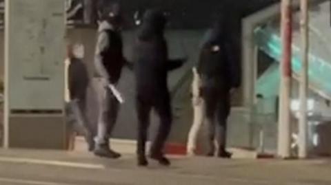 Three masked men and one is holding a machete