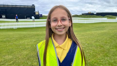 A young girl wears a yellow Hi-Viz vest. Underneath she wears a blue jumper and yellow polo shirt. Behind her is the back of a large navy stand. There is a green grass putting green over her right shoulder. 
