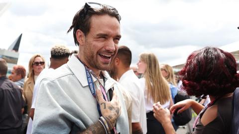 Liam Payne touches his chest as he smiles while wearing sunglasses and a white shirt with a crowd of people around him, during the Silverstone F1 Grand Prix in July 2023