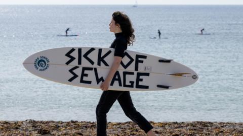 A female surfer carries a surfboard with the words "sick of sewage" written on it