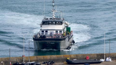 A large vessel with people in life jackets, arriving at a port. 