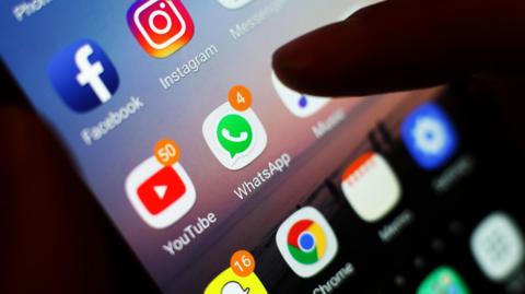 A mobile phone screen displaying several apps, with an index finger hovering over the WhatsApp icon.