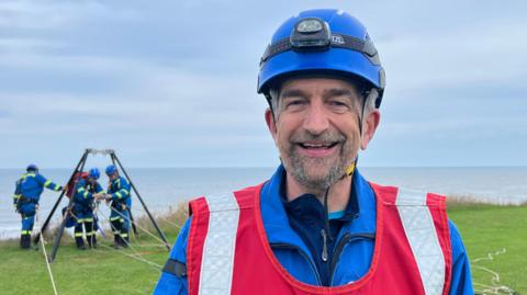 Jonathan Cliff standing on cliffs in Hornsea with a coastguard rescue team in the background he wears a blue uniform and helmet with a headtorch and a red safety vest.