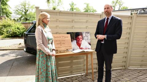 A woman with blonde hair wearing a long dress and light jacket stands near the prince who is wearing a suit and red tie and talking. Between them is a photo of a young man. There is also a gold plaque which says James' Place. 