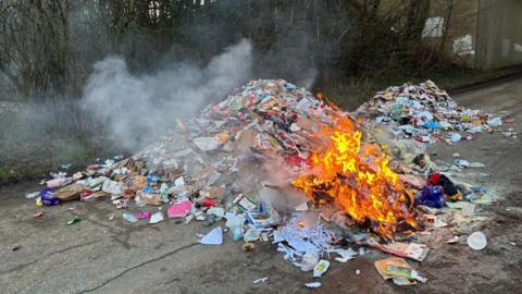 A large pile of rubbish dumped by the side of the road as a small fire burning on part of its surface. The rubbish is multi-coloured and largely appears to be discarded plastic