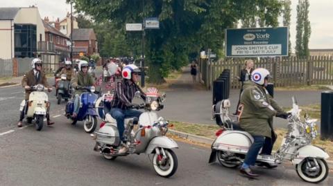In the foreground are four scooters - two silver, one dark blue and one cream coloured - turning into the entrance of York racecourse from a residential street. Three of the riders are wearing green fishtail parka coats and the other is wearing a brown and white striped long-sleeved shirt. Behind them can be seen a line of other scooters arriving along the street.