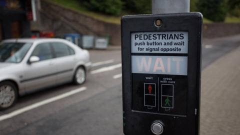 The push button of a pedestrian crossing with a grey saloon car driving past in the distance.