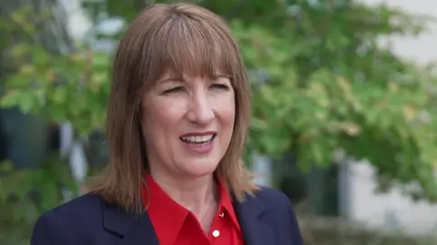 Rachel Reeves, wearing a red shirt and black suit, stand in front of a tree