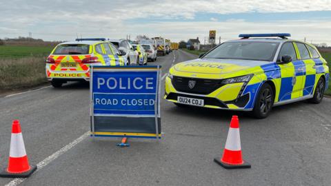 Emergency services are parked on the left side of the road and there is one police car on the right. There is a sign which says POLICE ROAD CLOSED in the middle.