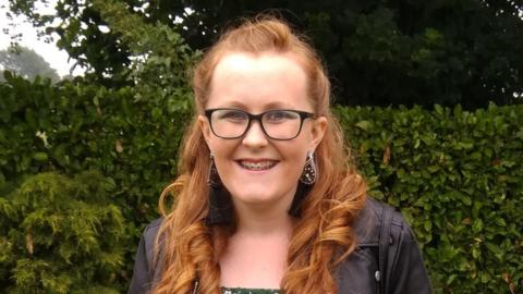 A head and shoulders shot of Amy Cooper stood in a garden, smiling at the camera. She has long, curled, ginger hair and is wearing black-rimmed glasses, silver, dangly earrings and a black leather jacket.