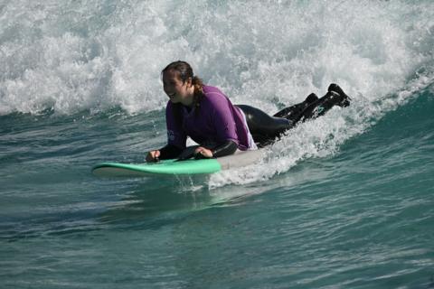 Kay Millar surfing on a green and grey surfboard. She is wearing a black wetsuit under a purple rash vest.