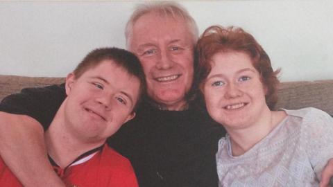Mark Jones sits in the middle of the sofa with his two adult children and he's wrapping their arms around them. His son - on the left - is wearing a red football shirt, and his daughter - on the right - is wearing a light patterned top