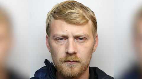 Man with fair hair and a beard and moustache narrows his eyes in a police mugshot.