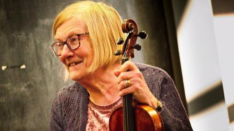 A woman with blond hair is smiling. She is holding a fiddle. She is wearing square-framed glasses and a grey cardigan. 