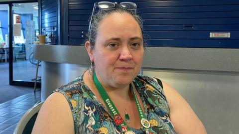 Becky Pain-Tolin sat down with a colourful dress and a green lanyard around her neck. She has glasses on her head. 