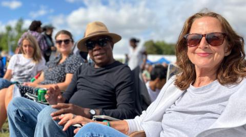 A man in a hat sits on a camp chair in a park, with one woman to his left and two to his right. The sun is shining, there are people in the background and festival food stalls