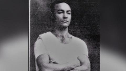A black and white picture of Sydney Bolesworth with his arms folded. He is wearing a white t shirt.