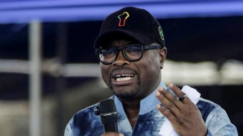 Benin s Finance Minister Romuald Wadagni in a blue shirt and a black cap, holding a microphone