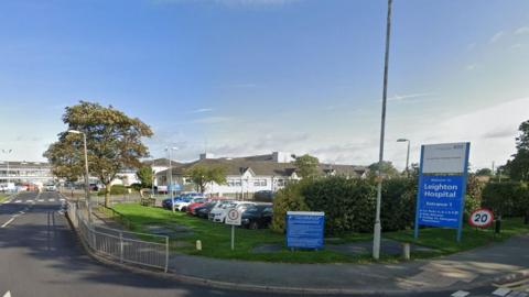 General view of the entrance to Leighton Hospital. A sign for the hospital is in the foreground along with a pavement and car park, the hospital building is visible in the background