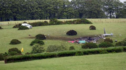 Scene of a helicopter crash in a field. Debris from the grey military helicopter is strewn across the grass. Wreckage can be seen across parts of the field. Tarpaulin is on top of the wreck.