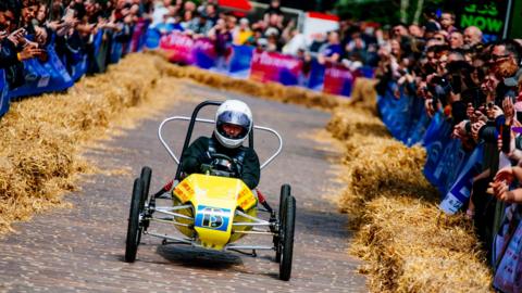 A go-kart with a driver wearing a crash helmet drives along a cobbled street with spectators either side behind hay bales.