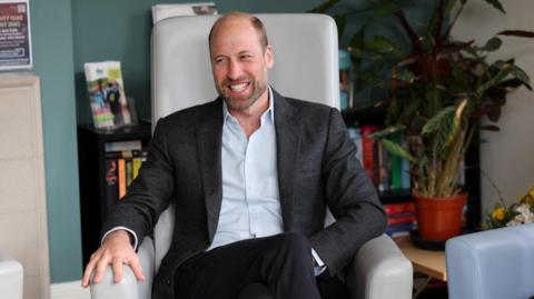 Prince William smiling at the camera and sitting in an armchair. He is wearing a dark suit and pale shirt.