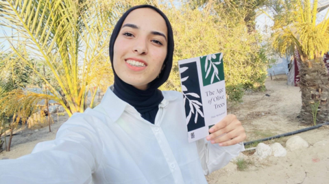 Haia Mohammed, wearing a white shirt and black headscarf, smiles while holding a book titled The Age of Olive Trees in an outdoor setting with palm trees in the background.