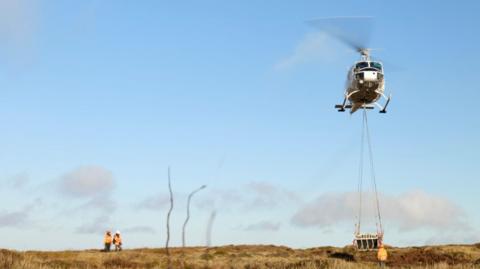 A helicopter hovering above moorland with supplies hanging beneath it