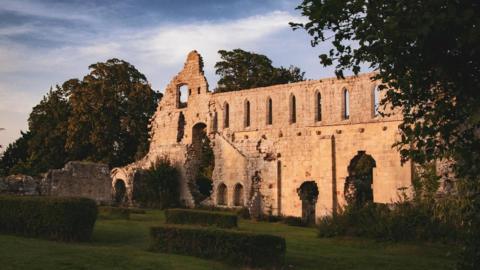 A stone abbey which is in ruins set in gardens with neatly-cut hedges. 