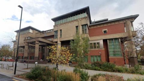 Exterior of Sheffield Crown Court, a red brick and stone building with columns and windows. In the foreground are trees and bushes.