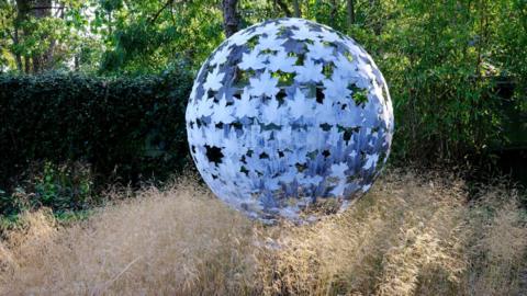 A blue sphere sculpture that appears to be made of large leaves sits in a long grassy area in front of a dark green hedge