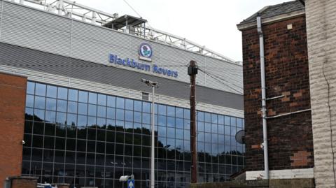 The outside of Ewood Park, showing a glass-fronted stand with the club logo of a red rose surrounded by a blue ring.