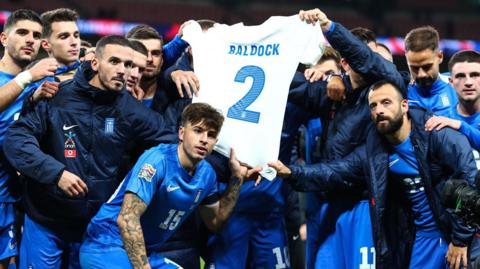 Greece players hold up a shirt bearing George Baldock's name and number two after their victory against England