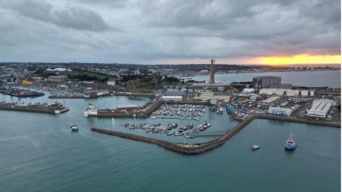 A drone picture of a harbour in Jersey. The water is blue and the sunset can be seen in the distance.