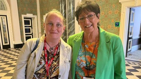 A woman in a cream jacket wearing a red lanyard that says visitor stands next to another woman who is wearing a green blazer. They are in a council building, which has black and white chequered floors and floral wallpaper.