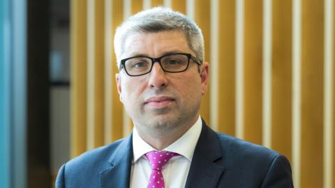 A man with short hair and glasses poses for a portrait picture. He is standing in an office setting and wearing a navy suit, white shirt and pink tie with white spots.