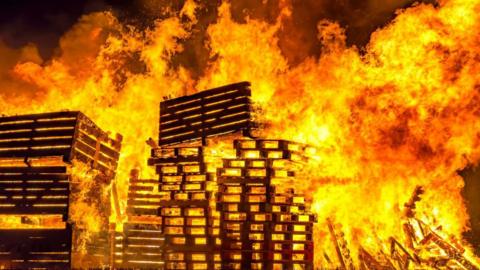 A large fire with wooden pallets visible in the flames